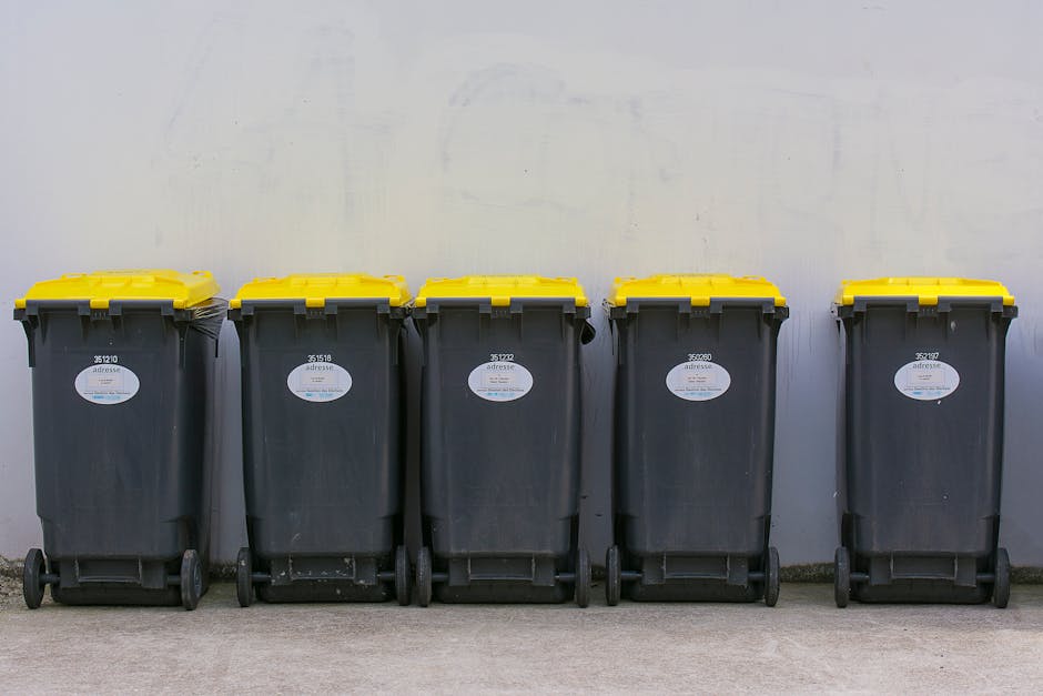 A collection of overflowing rubbish bins and scattered waste bags and cardboard boxes is situated on a paved area in front of a commercial building. The central bin is a large, grey, mixed paper and cardboard container with its lid partially open, revealing crumpled paper, flattened cardboard, and various paper materials inside. Surrounding the central bin are black and red waste bags, flattened cardboard boxes, and loose trash, including paper, plastic packaging, and other debris. To the left, a small silver car is parked alongside a metal railing, with a tree behind it. The background features the facade of a building under construction or renovation, covered in blue scaffolding and protective sheeting. The scene appears to be in an urban environment, possibly during daylight, in an area designated for waste collection, illustrating a typical scenario of rubbish accumulation awaiting collection or disposal services by waste management companies such as Waste Collection Wimbledon.