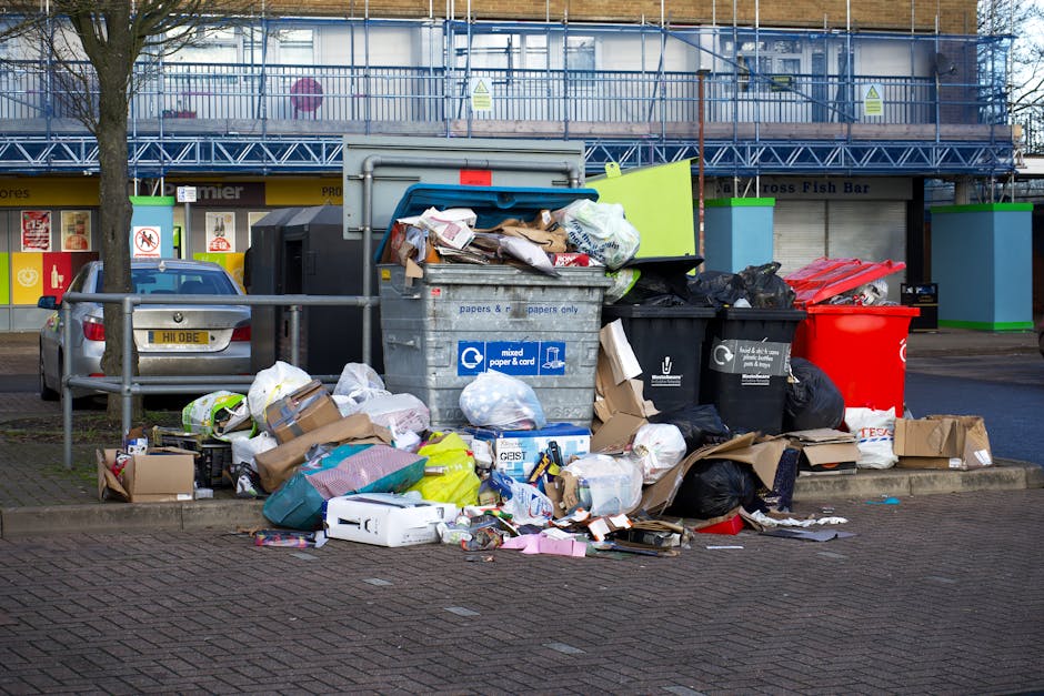 A large outdoor waste collection area showing multiple overflowing black and black plastic rubbish bags, cardboard boxes, and paper waste piled on and around a metal recycling bin labeled for mixed paper and card. The black bins are partially open, revealing some debris inside, while a bright red wheelie bin is positioned nearby. Behind the waste, there is a sidewalk with a metal railing and a parked silver car, and in the background, a building with a blue scaffolding structure covering the upper floor and a row of shops with brightly coloured signage. The scene appears to be in a commercial or urban area, reflecting a temporary or unmanaged rubbish accumulation that might require professional disposal services, such as those offered by Waste Collection Wimbledon, to support private waste handling or alternative collection arrangements.