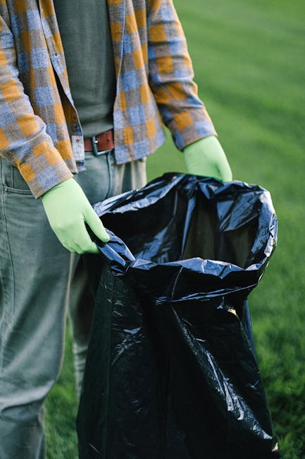 A person wearing a plaid shirt with yellow and blue tones, a green long-sleeve shirt, and light grey trousers, is standing outdoors on a grassy area. They are holding open a large black plastic rubbish bag with both hands, which is partially lined with a grey interior. The person's hands are covered with light green rubber gloves, indicating they are engaged in waste collection or disposal. The background features a well-maintained green lawn, suggesting an outdoor or garden environment. The scene emphasizes the act of private waste handling or rubbish removal, consistent with services offered by Waste Collection Wimbledon. The lighting appears natural, highlighting the textures of the plastic bag, fabric, and grass, supporting a professional and factual visual description of an on-site waste collection activity.