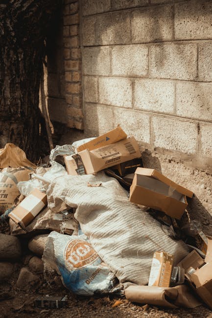 A large outdoor waste collection area showing multiple overflowing black and black plastic rubbish bags, cardboard boxes, and paper waste piled on and around a metal recycling bin labeled for mixed paper and card. The black bins are partially open, revealing some debris inside, while a bright red wheelie bin is positioned nearby. Behind the waste, there is a sidewalk with a metal railing and a parked silver car, and in the background, a building with a blue scaffolding structure covering the upper floor and a row of shops with brightly coloured signage. The scene appears to be in a commercial or urban area, reflecting a temporary or unmanaged rubbish accumulation that might require professional disposal services, such as those offered by Waste Collection Wimbledon, to support private waste handling or alternative collection arrangements.