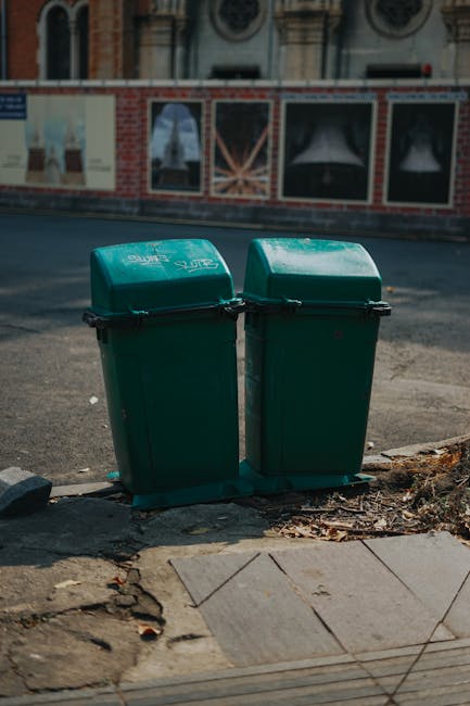 Two green plastic waste bins with black lids are positioned side by side on a paved sidewalk, close to the edge of a road. The bins are slightly tapered, with a smooth finish and locked lids, and appear to be used for general rubbish collection. They are situated on a concrete curb, with some fallen leaves and small debris around their base. In the background, there is a brick wall with large, transparent window panels, behind which a building with decorative architectural features is partially visible. The scene is outdoors, illuminated by natural daylight, and the road surface behind the bins appears clean but shows typical urban wear with some gravel and minor stains. The overall environment suggests an area where private waste disposal, managed by waste collection services like Waste Collection Wimbledon, takes place, offering an alternative to local authority rubbish removal for residents or businesses in the area.